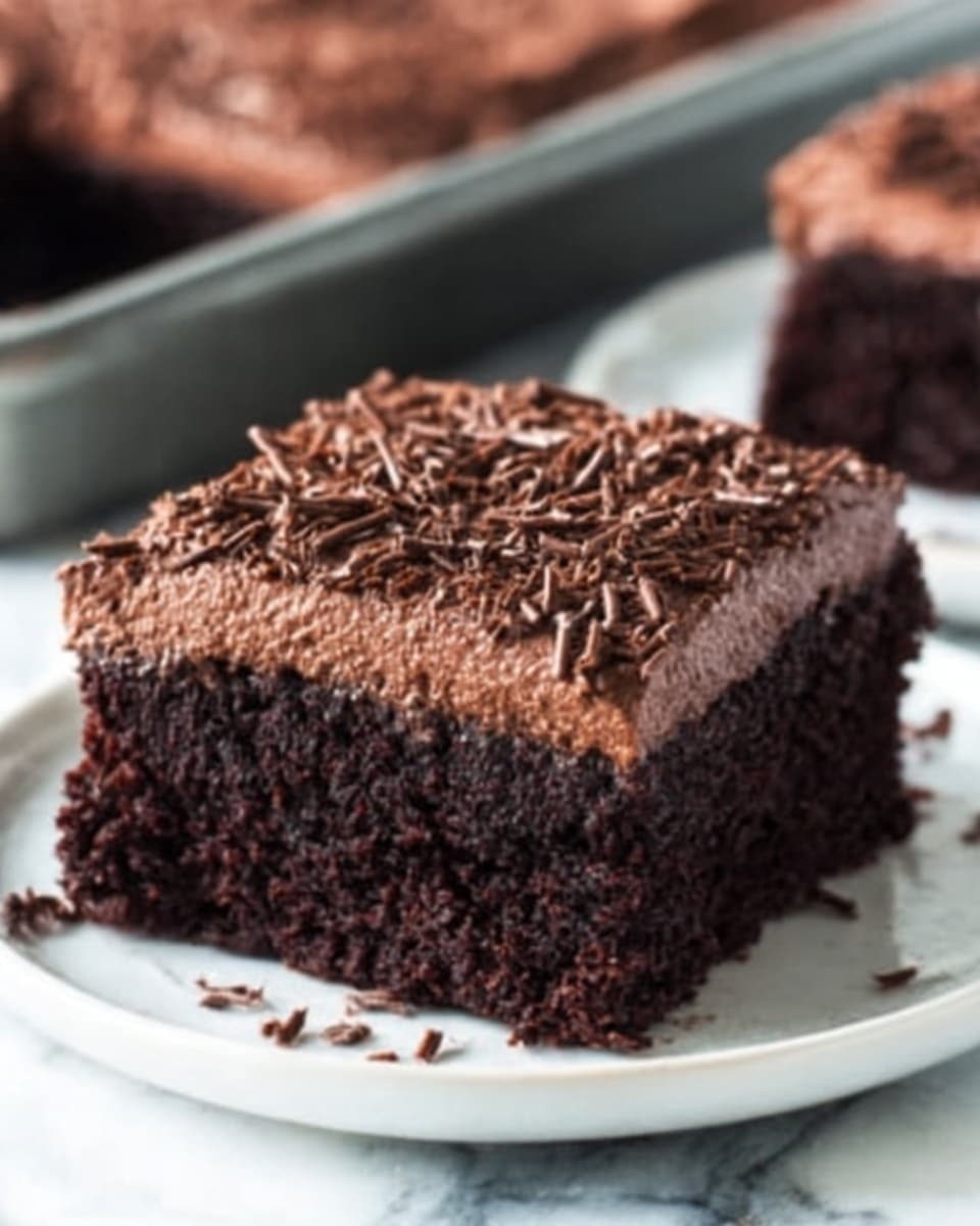 A close-up image of a single square piece of moist chocolate cake on a white plate. The cake has two layers: a rich, dark brown chocolate base with a slightly crumbly texture, and a thick, smooth layer of light brown chocolate frosting on top. There are fine chocolate shavings scattered on the frosting, adding a delicate texture. The plate sits on a white marbled surface, and in the background, there is a blurred baking pan with more cake and a small white bowl filled with chocolate chips. Photo taken with an iphone --ar 4:5 --v 7