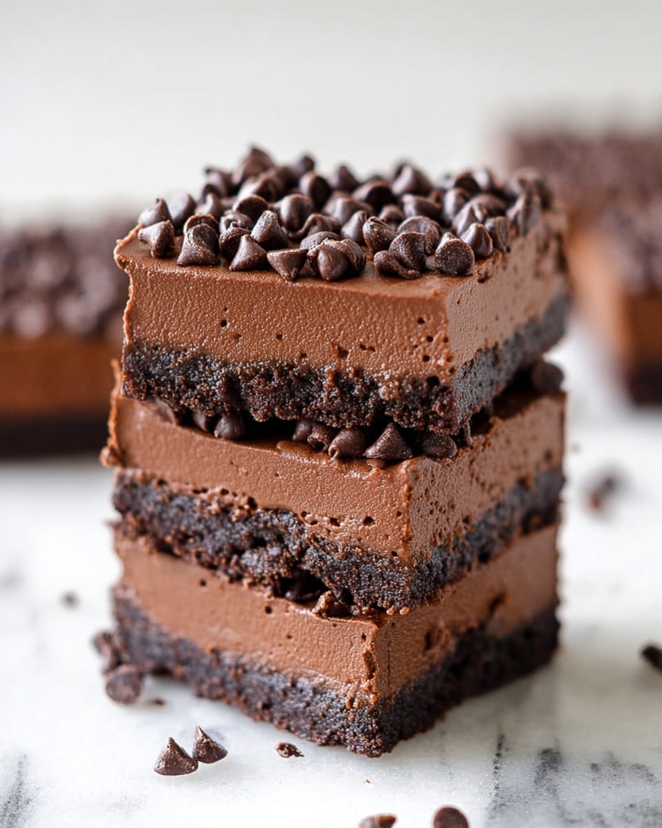 Three square chocolate dessert bars are stacked on top of each other on a white marbled surface, each bar showing two layers: a dark, crumbly base that is thin but firm, and a thick, smooth chocolate layer above it with rich texture. The top layer is generously covered with small, shiny chocolate chips that add a bumpy texture and darker spots throughout. Around the stack, more pieces of the same dessert can be seen blurred in the background. The overall look is dense, creamy, and chocolatey with a contrast between the crumbly base and smooth middle. photo taken with an iphone --ar 4:5 --v 7