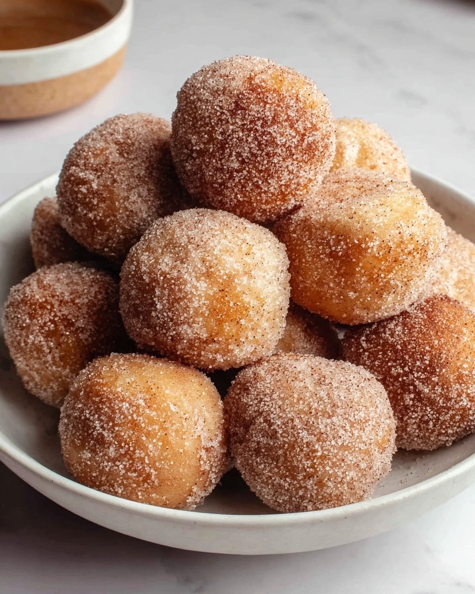 A white bowl filled with a pile of round doughnut holes, each coated in a mix of sugar and cinnamon, giving them a speckled light brown and white texture. The doughnut holes are closely packed, showing their soft and slightly rough surface from the sugar coating. The bowl sits on a white marbled surface with a blurred bowl in the background. photo taken with an iphone --ar 4:5 --v 7