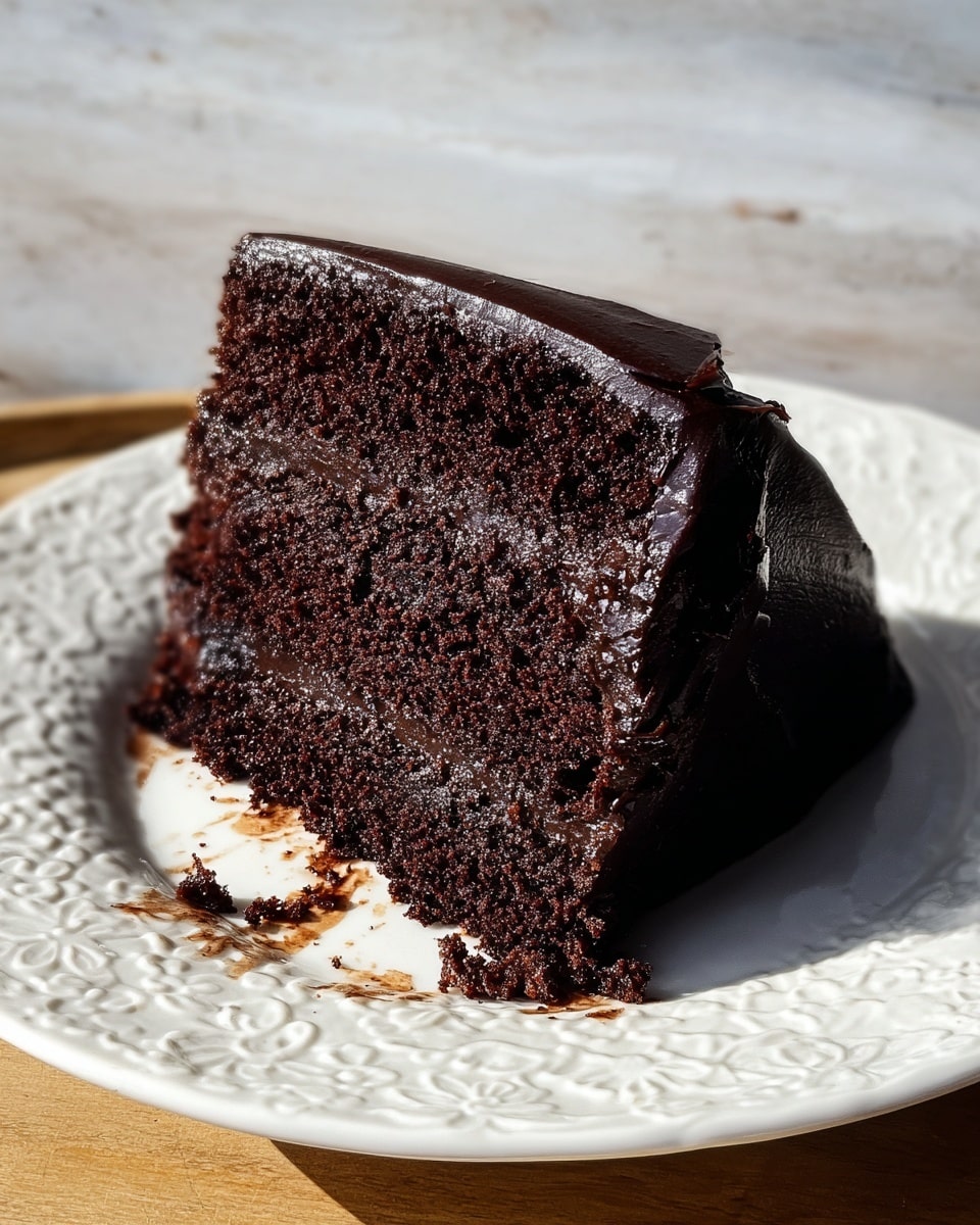 A slice of dark chocolate cake with three layers of moist, rich, almost black cake separated by two layers of smooth, dark chocolate frosting. The outside of the cake is coated with shiny, thick chocolate icing with a few small crumbs pressed into the sides. The slice sits on a white plate with a delicate raised pattern, and a bit of smeared chocolate is visible on the plate beneath the cake. The scene is lit with warm sunlight, casting soft shadows and bringing out the deep textures of the cake. Photo taken with an iphone --ar 4:5 --v 7