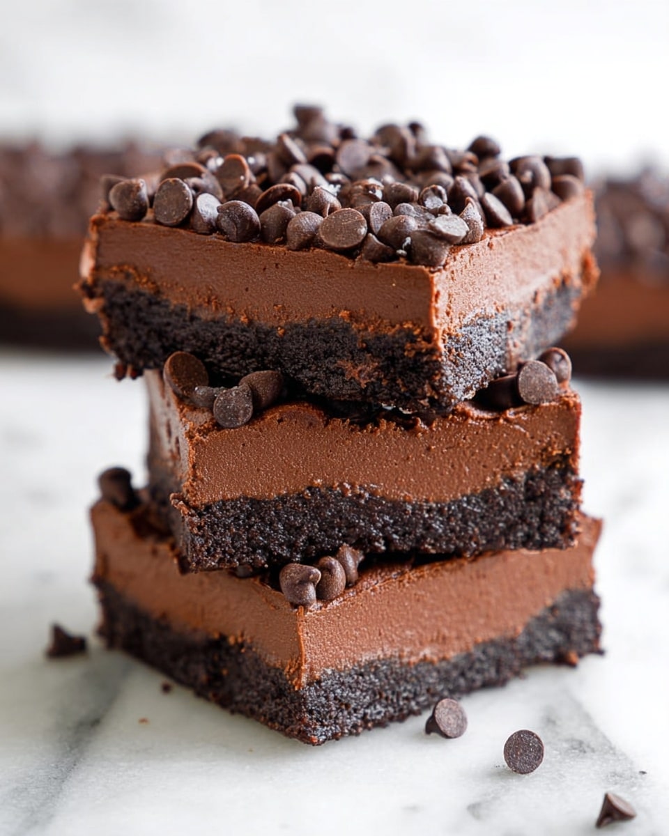 The image shows a stack of three chocolate dessert squares on a white marbled surface. Each square has two layers: a dark, crumbly bottom crust and a thick, smooth, creamy chocolate layer on top. The chocolate layer is rich and dense, covered with many small, shiny chocolate chips scattered across the surface, adding texture and visual interest. A small part of another piece is visible on the sides, blurred in the background. Photo taken with an iphone --ar 4:5 --v 7
