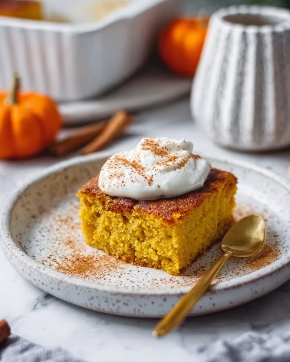 A square piece of golden pumpkin cake with a moist texture sits in the center of a white plate with subtle speckles. On top of the cake is a thick dollop of white cream, sprinkled lightly with cinnamon powder. A gold spoon lies next to the cake on the plate. The setting is on a white marbled surface, with a white textured vase and a small pumpkin in the blurred background, giving a cozy, autumn feeling. Photo taken with an iphone --ar 4:5 --v 7
