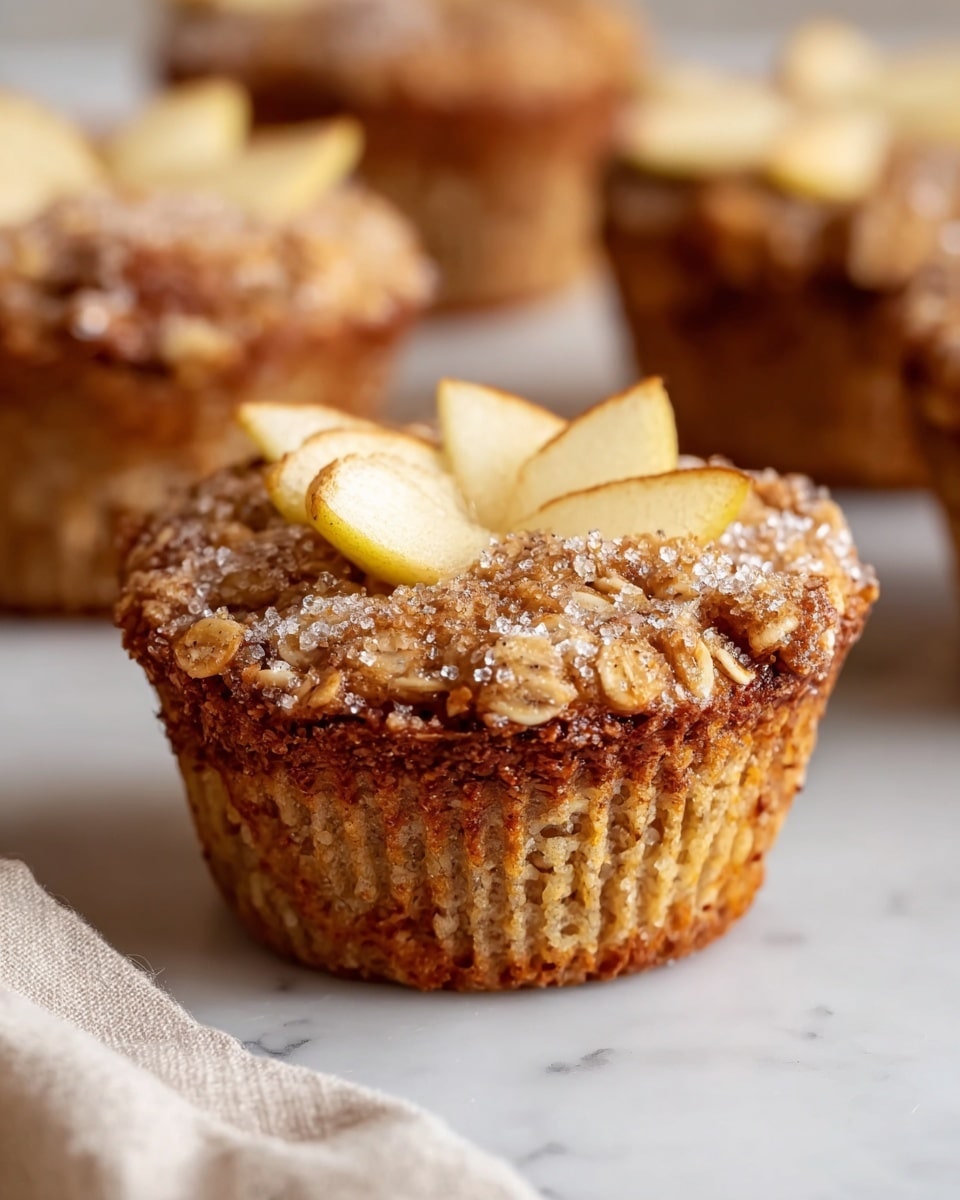 The image shows a close-up of a single muffin with visible layers and textures. The bottom layer is a light brown baked muffin base with a slightly rough, crumbly texture. The middle layer is topped with a mixture of oats and small slices of apple, giving it a chunky and rustic look with light and golden brown colors. The top layer features several apple slices partially embedded in the oat mixture, sprinkled with coarse sugar crystals and a dusting of cinnamon powder, adding a sparkly and speckled texture. The muffin is placed on a white marbled surface with a soft cloth nearby, and two blurred muffins are visible in the background. photo taken with an iphone --ar 4:5 --v 7