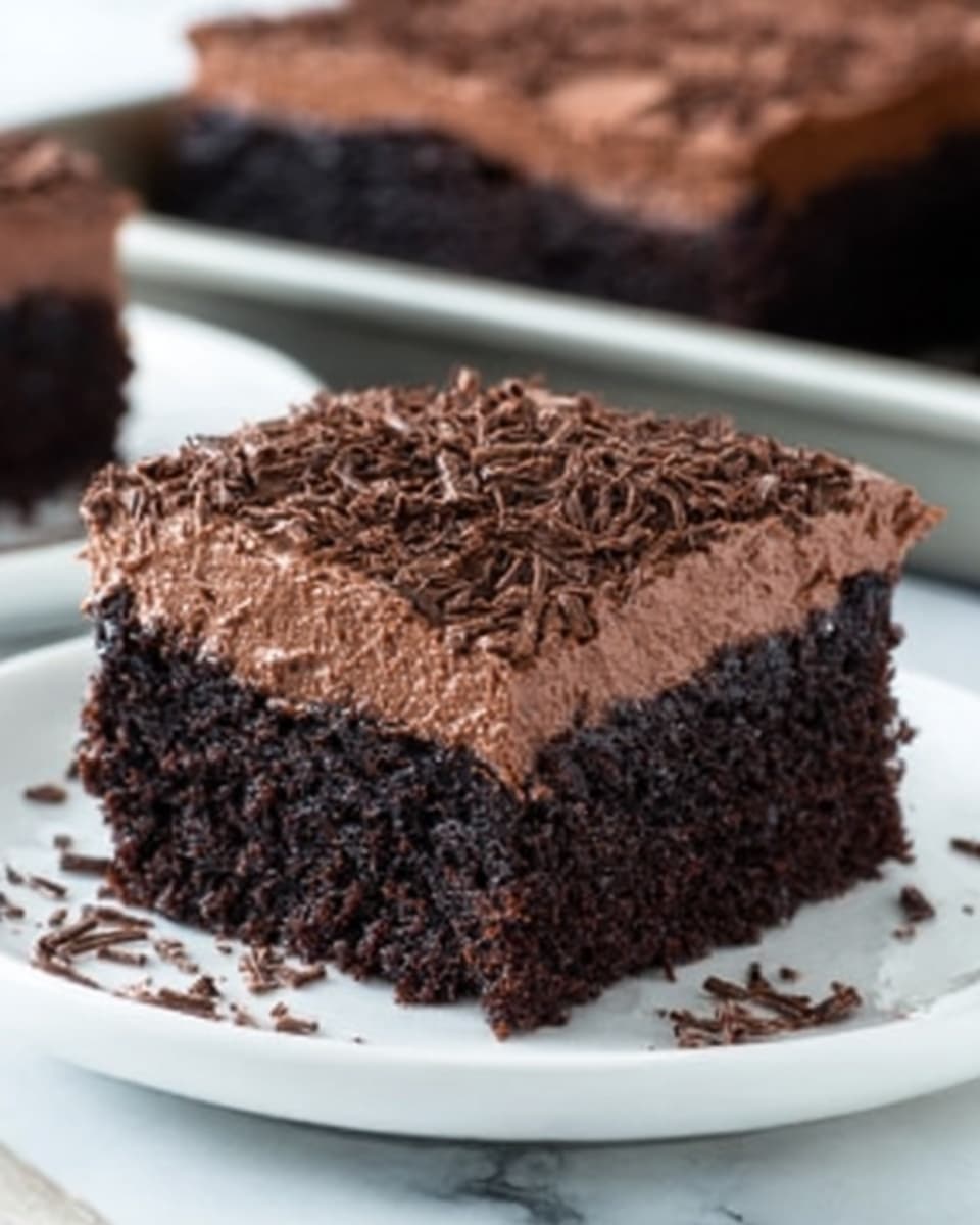 A square piece of dark chocolate cake with a thick layer of smooth, creamy chocolate frosting on top. The top of the frosting is sprinkled with thin chocolate shavings, adding a bit of texture. The cake looks moist and dense with tiny air holes showing inside. It is placed on a white plate with a simple design. The background shows a white marbled surface, and there is a blurred pan of similar cake pieces in the back. photo taken with an iphone --ar 4:5 --v 7