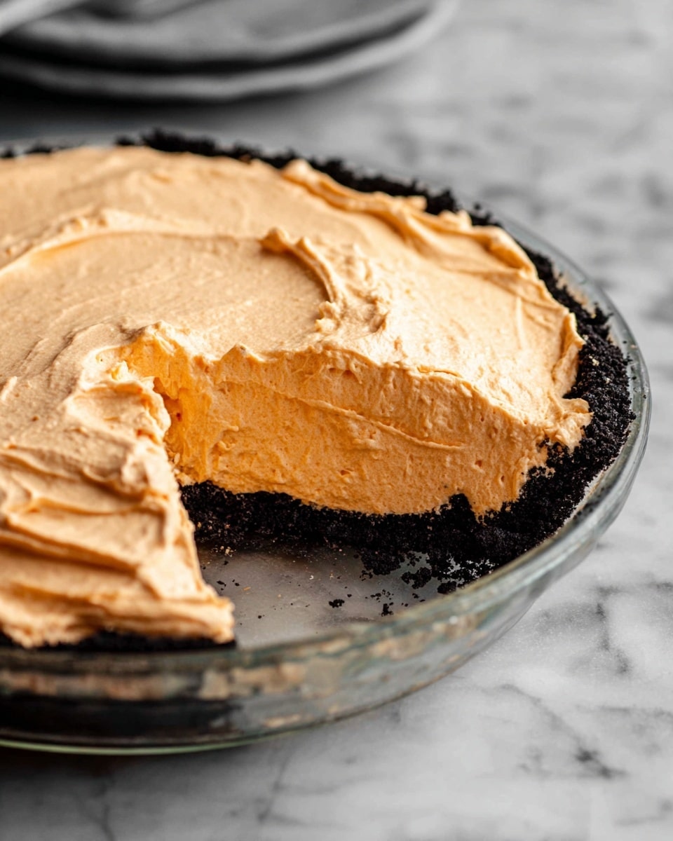 The image shows a three-layer pie in a clear glass dish sitting on a white marbled surface. The bottom layer is a dark, crumbly crust, almost black in color, forming a solid base. The middle layer is a thick, smooth, light orange filling, with a creamy texture that looks soft and fluffy. The top layer is the same light orange color, whipped smoothly with some swirled patterns, giving it a light and airy appearance. A slice has been cut out from the pie, showing the clean layers clearly from the side. Photo taken with an iphone --ar 4:5 --v 7