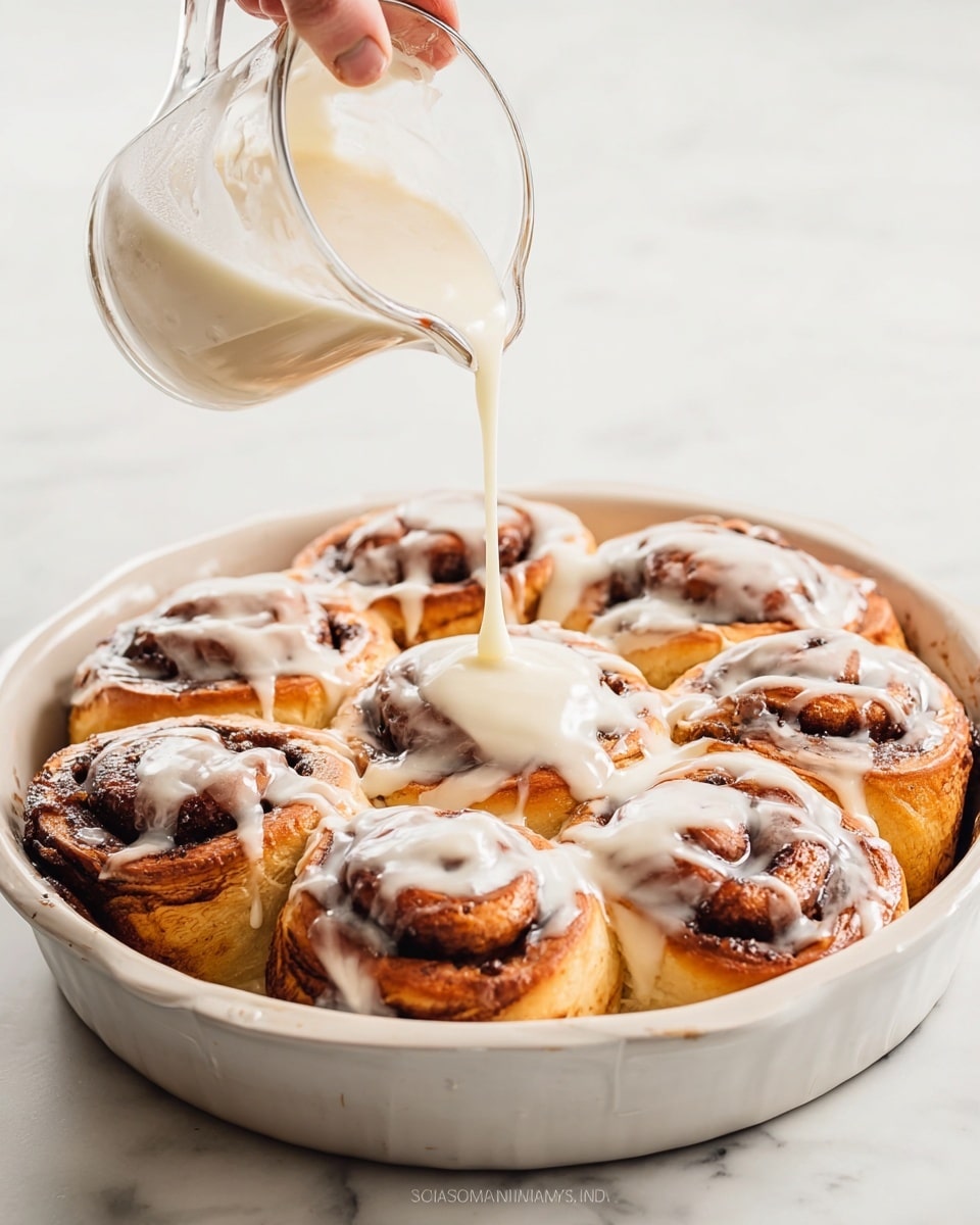 A white scalloped ceramic dish holds nine golden-brown cinnamon rolls, each with dark brown swirls of cinnamon filling visible in the spiral layers. The rolls are freshly baked, puffy and soft with a light crust around the edges. A woman's hand pours a creamy white glaze from a small clear glass pitcher, drizzling it over the rolls, creating a shiny, smooth layer that slowly spreads and drips down the sides of each cinnamon roll. The scene is set on a white marbled surface with a clean, bright background. photo taken with an iphone --ar 4:5 --v 7