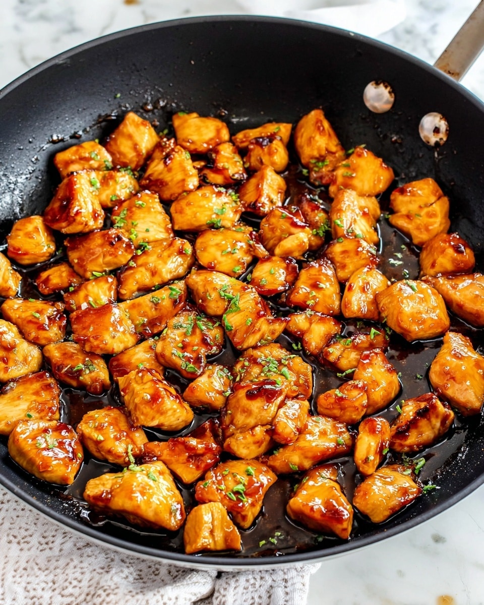 A black pan filled with many small, cooked chicken pieces coated in shiny, brown sauce. The chicken pieces are golden-brown with slight caramelized marks and scattered small green herb bits on top. The pan is placed on a white marbled surface with a light brown cloth partly visible underneath. The overall look is warm and appetizing with a glossy texture on the chicken. Photo taken with an iphone --ar 4:5 --v 7