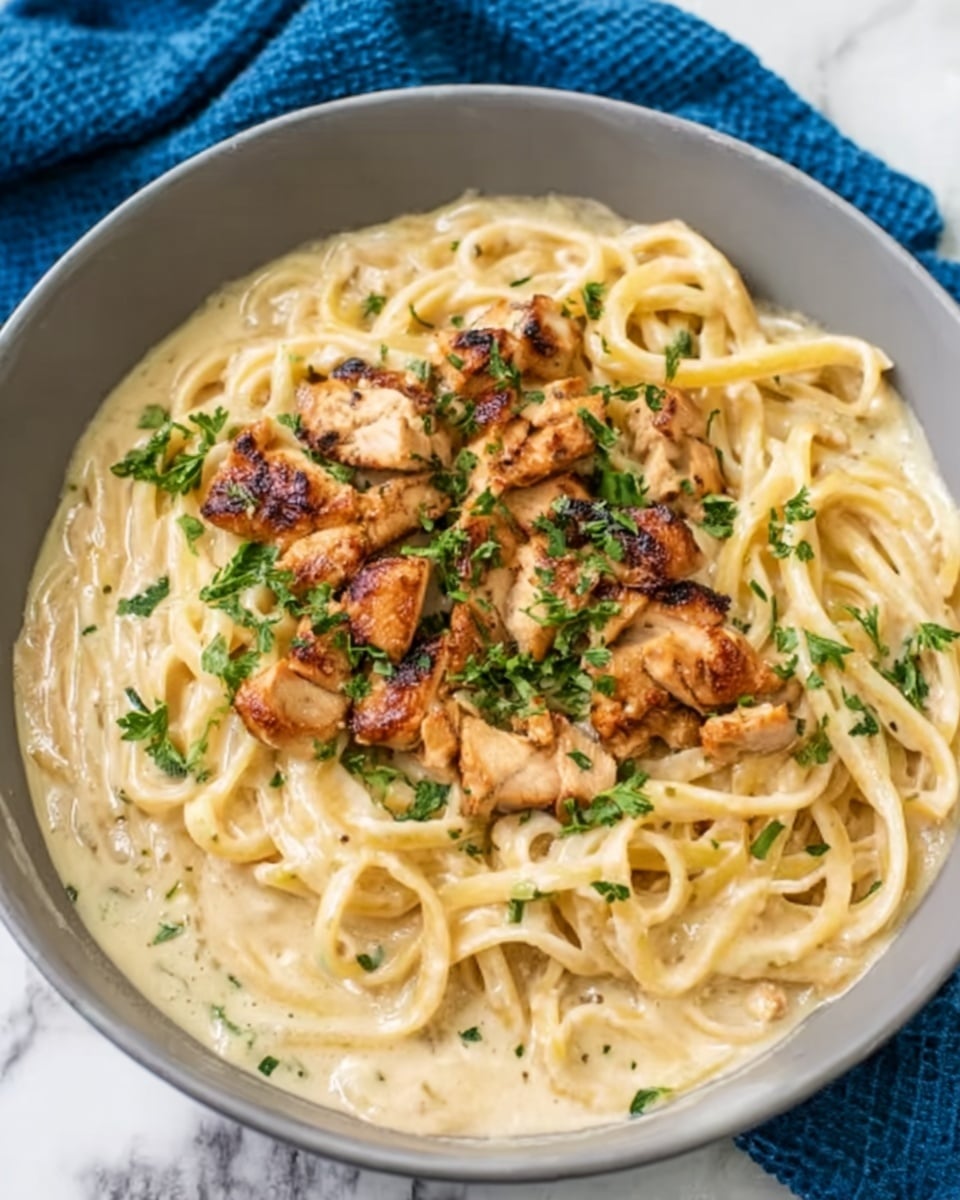A white bowl holds a creamy pasta dish with thick spaghetti noodles fully covered in a smooth, light beige sauce. On top, there are small pieces of browned grilled chicken scattered in the center, adding a golden-brown contrast. Fresh green herbs are sprinkled lightly over the chicken and sauce, giving a fresh touch. The bowl sits on a white marbled surface with a blue cloth partially visible at the side. Photo taken with an iphone --ar 4:5 --v 7