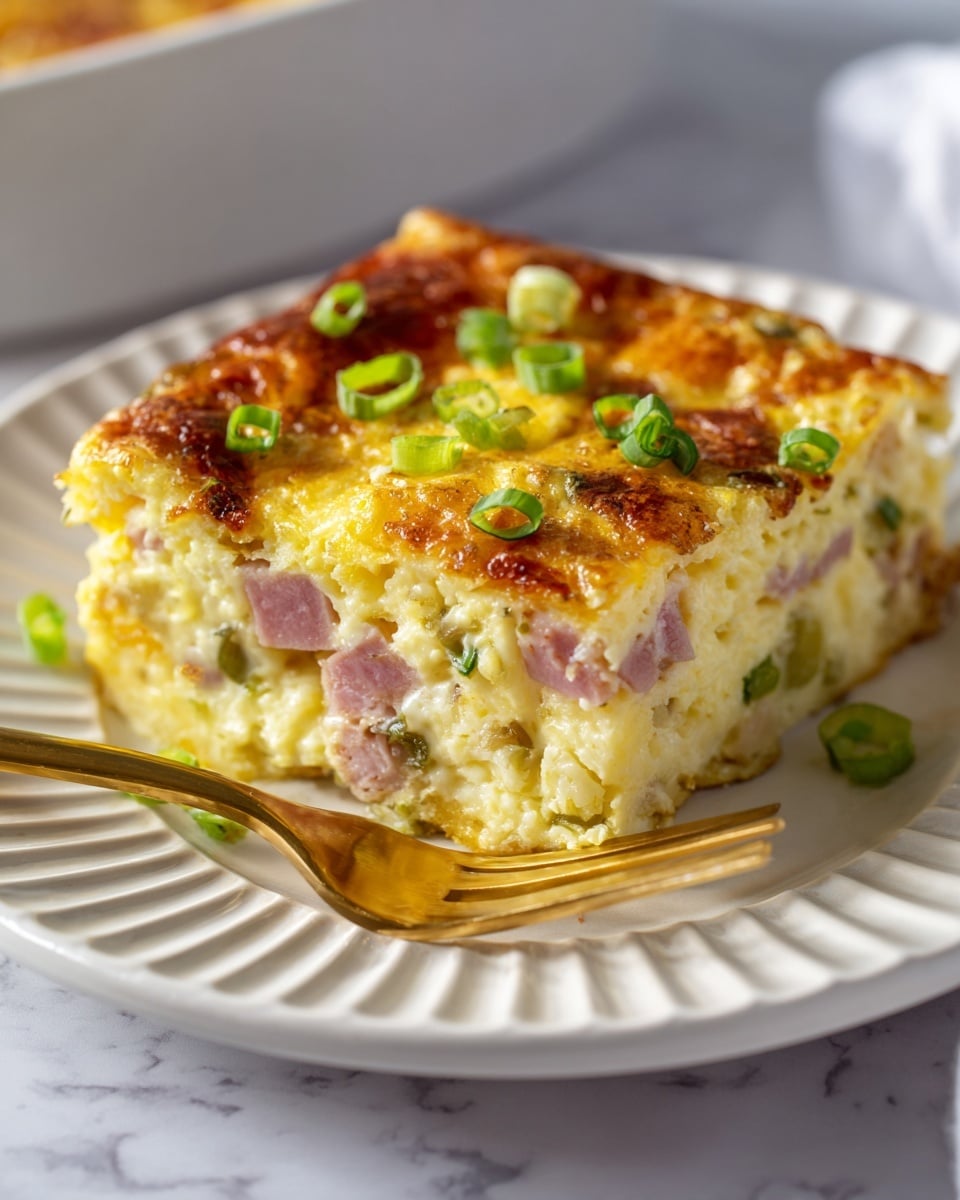 A square slice of baked egg casserole sits on a white ridged plate with a golden fork next to it. The casserole has three visible layers: a firm golden brown top with small browned spots and topped with three thin rings of green onion, a thick creamy middle filled with a mix of light yellow eggs, diced pink ham, and green bits of herbs or vegetables, and a bottom layer that is soft and pale yellow with a slightly browned edge. The white marbled background adds a clean bright look to the image. Photo taken with an iphone --ar 4:5 --v 7