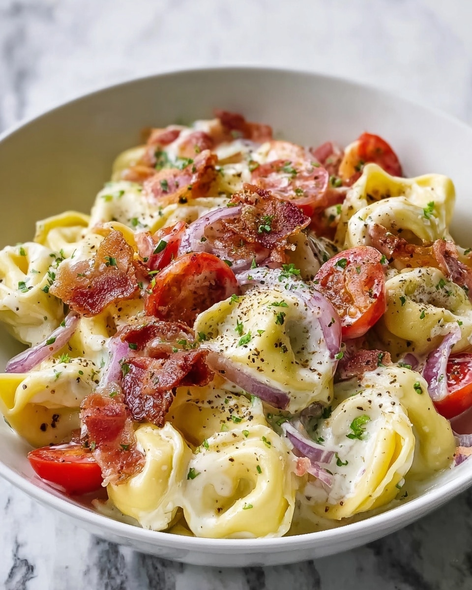 A white bowl is filled with creamy tortellini pasta, each piece yellow and plump with a smooth texture. Mixed in are bright red cherry tomato halves and thin slices of purple onion, adding pops of color. Crispy brown bacon pieces are scattered throughout the dish. A creamy white sauce coats the pasta, sprinkled with finely chopped green herbs and black pepper for texture and detail. The bowl sits on a soft gray cloth, all placed on a white marbled surface. Photo taken with an iphone --ar 4:5 --v 7