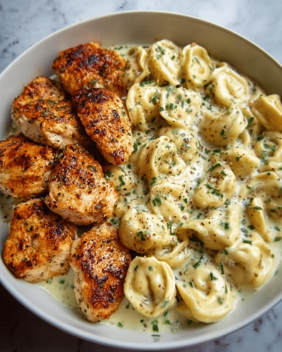 The image shows a white bowl filled with two main parts: on the left, several pieces of cooked chicken with a golden-brown crispy texture and light seasoning, and on the right, creamy tortellini pasta coated in a rich, white sauce with herbs sprinkled on top. The sauce looks thick and smooth, with small green herb bits scattered around both the chicken and pasta. The dish sits on a white marbled surface, creating a clean and bright background. Photo taken with an iphone --ar 4:5 --v 7