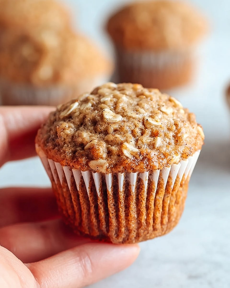 A close-up image of a single oatmeal muffin held gently by a woman's hand, showing fine texture with visible oats sprinkled on top and a golden-brown crust. The muffin has a slightly domed top that looks soft and moist with small specks of cinnamon or spice giving it a rustic appearance. The muffin liner is white with light ridges wrapping around the base. In the blurred background, there are more muffins resting on a white marbled surface. photo taken with an iphone --ar 4:5 --v 7