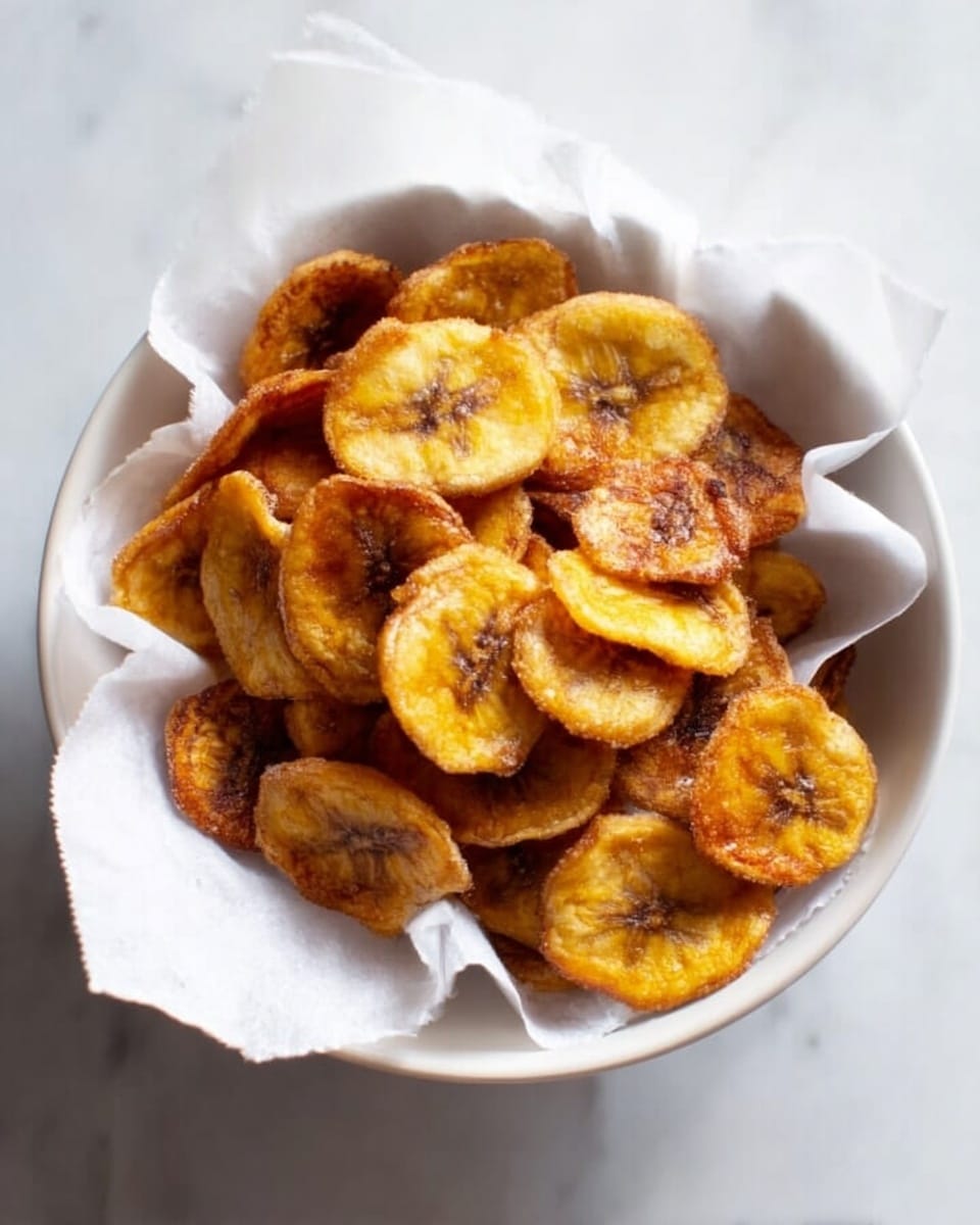 A white bowl lined with crumpled white paper holds several golden-brown plantain chips, each chip showing a slightly crispy texture with small darker spots of caramelization. The chips are thin and round, layered loosely with some overlapping inside the bowl. The background features a white marbled surface that adds a clean and simple look to the scene. Photo taken with an iphone --ar 4:5 --v 7
