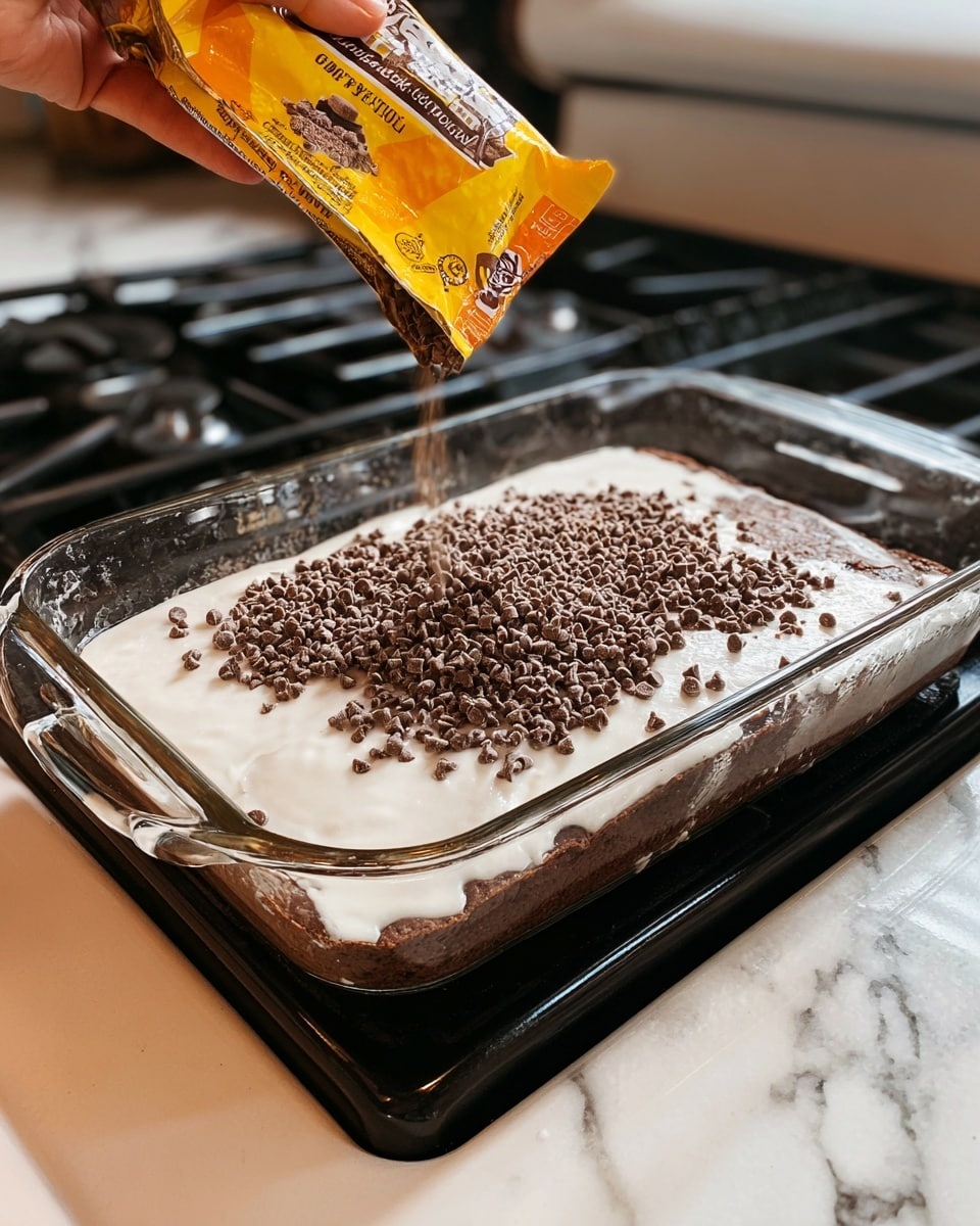 A clear glass rectangular baking dish holds a dessert with two main visible layers. The bottom layer is a moist, dark brown cake. On top, a thin shiny layer of white creamy glaze evenly covers the cake. A woman's hand is shown pouring small, dark brown chocolate chips from a yellow and orange package onto the glaze, creating a scattered textured top layer. The dish is set on a white marbled stove top near black electric coil burners. photo taken with an iphone --ar 4:5 --v 7