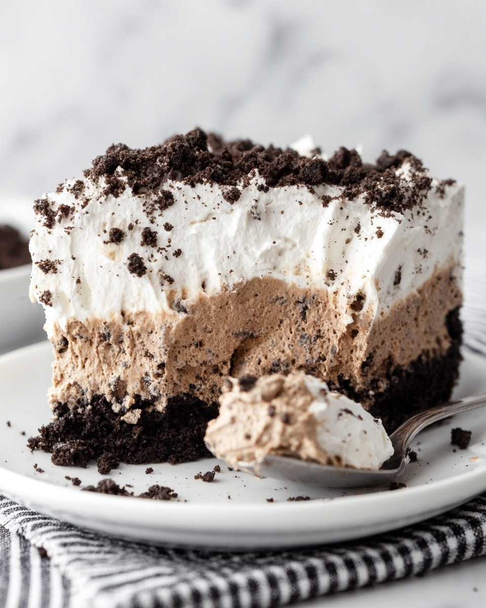 The image shows a close-up of a thick slice of creamy layered dessert on a white plate with a black and white striped cloth underneath. The dessert has three distinct layers: the bottom is a dark, crumbly chocolate crust, the middle is a light brown mousse-like layer with small dark cookie bits mixed in, and the top is a white whipped cream layer sprinkled with more dark cookie crumbs. A spoon on the right side holds a scoop of the middle and bottom layers, showing the soft, airy texture of the mousse. The background is a white marbled texture. photo taken with an iphone --ar 4:5 --v 7
