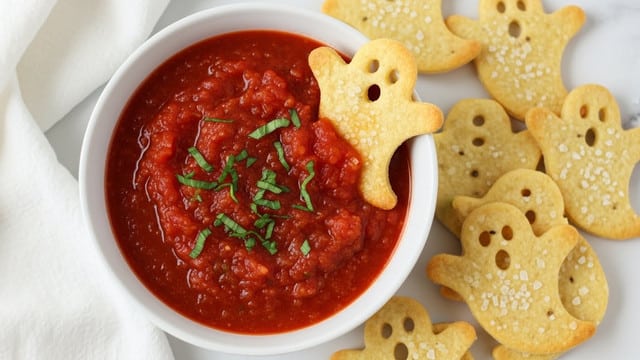 The image shows many ghost-shaped tortilla chips scattered on a surface with a white marbled texture. Each chip is light golden with some darker spots and has visible coarse salt sprinkled on top. In the center, there is a small white bowl filled with chunky red salsa that has pieces of green cilantro on it. One tortilla chip is partially dipped into the salsa in the bowl. A white cloth is partially visible in the top left corner of the image. The overall look is bright and festive. photo taken with an iphone --ar 4:5 --v 7