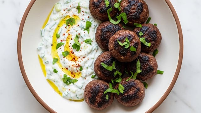 A white shallow bowl holds nine dark brown grilled meatballs with a slightly crispy texture, scattered on the right side of the bowl. On the left side, there is a creamy white sauce with green herb pieces mixed in, spread out in a loose crescent shape. Bright green chopped parsley is sprinkled over both the meatballs and the sauce, adding color contrast. There is also a light dusting of red spice powder over the sauce and some parsley. The bowl sits on a white marbled surface. Photo taken with an iphone --ar 4:5 --v 7