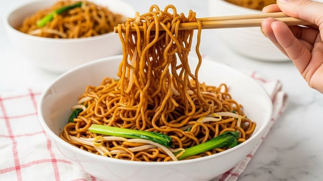 A white bowl filled with dark brown noodles mixed with light beige bean sprouts and green vegetable pieces. The noodles look shiny and slightly oily, resting as the main layer in the bowl. A woman's hand uses light brown chopsticks to lift a thick portion of the noodles, showing their thin, curly texture. The bowl sits on a white marbled surface with a pink and white checkered cloth in the front and another white bowl with more noodles blurred in the background. Photo taken with an iphone --ar 4:5 --v 7