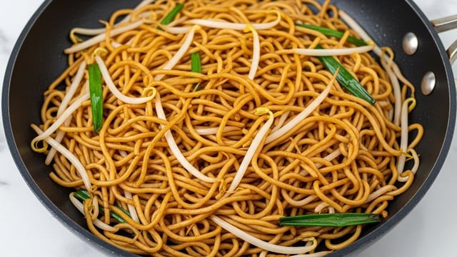 The image shows a close-up of stir-fried noodles in a black non-stick pan. The dish has one main layer of thin, golden-brown noodles tangled together, mixed with long white bean sprouts and some chopped green onions scattered throughout. The noodles have a slightly shiny texture from the sauce, and the bean sprouts add a crisp, smooth contrast. The pan is placed on a white marbled surface. photo taken with an iphone --ar 4:5 --v 7