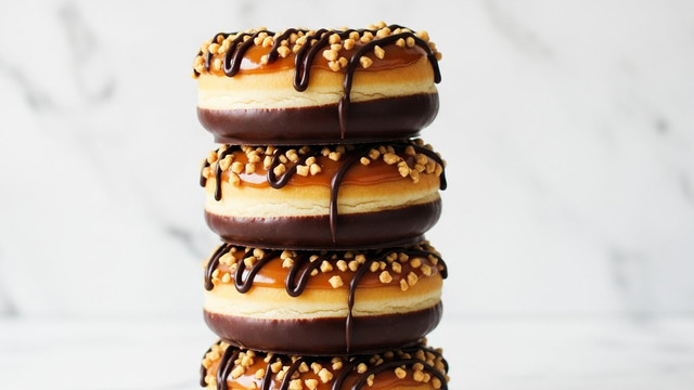 A tall stack of five round chocolate and caramel donuts is shown against a white marbled texture background. Each donut has a bottom layer coated in dark chocolate, with the middle section being a light tan donut dough. The top layer is covered with a shiny caramel glaze sprinkled with small, crunchy nut pieces. Thin dark chocolate lines are drizzled over the caramel and nuts on each donut, creating a textured pattern. The donuts are perfectly stacked, showing the alternating colors and textures clearly. Photo taken with an iphone --ar 4:5 --v 7