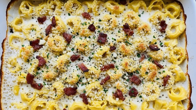 A white baking dish filled with a creamy baked tortellini dish, showing one layer of golden yellow tortellini pasta covered in a thick white cheese sauce. The top is sprinkled with small browned bits of crispy bacon and a light layer of golden toasted breadcrumbs. Small specks of green herbs and black pepper are scattered across the surface. The edges of the pasta are slightly browned from baking, with a bubbling cream sauce visible around the edges. Photo taken with an iphone --ar 4:5 --v 7