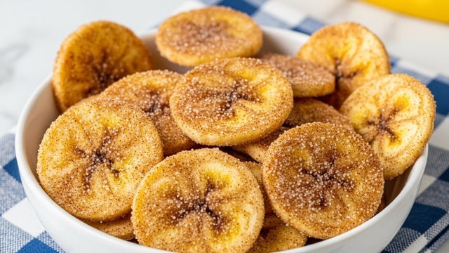 The image shows a white bowl filled with several thin, round banana chips that have a light golden-yellow color. Each chip is coated with a fine layer of brown cinnamon and sugar, giving them a textured, slightly sparkling surface. The chips are stacked loosely, some overlapping each other with a few chips standing upright at the edges. The bowl sits on a blue and white checked cloth against a white marbled surface with a hint of yellow in the background. photo taken with an iphone --ar 4:5 --v 7