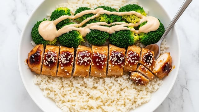 A white bowl holds a dish with three layers: the bottom layer is fluffy white rice, soft and evenly spread; the middle layer consists of vibrant green broccoli florets tenderly cooked, mixed with bite-sized pieces of golden-brown grilled chicken coated in a glossy dark sauce, arranged side by side; the top layer is a light cream sauce drizzled over the broccoli and chicken, with some sprinkled white sesame seeds for texture. A silver fork is partially inserted into the chicken. The bowl sits on a white marbled textured surface. photo taken with an iphone --ar 4:5 --v 7